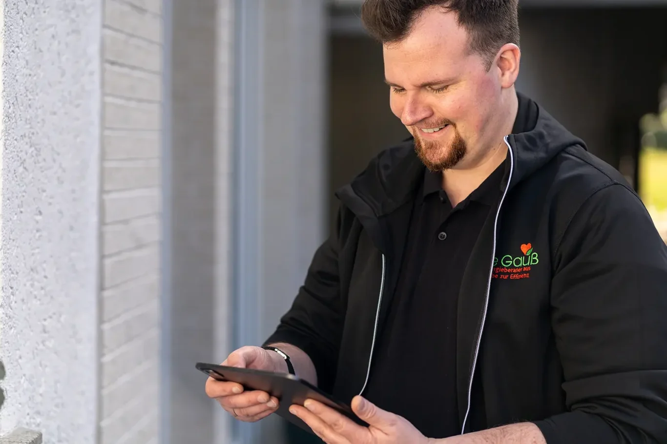 Dennis Gauß – Ihr zertifizierter Energieberater für Mainz & Umgebung Dennis Gauß, Inhaber von Energieberater De Gauß in Mainz, steht sympathisch lächelnd in schwarzer Arbeitsjacke mit Firmenlogo in einem moder