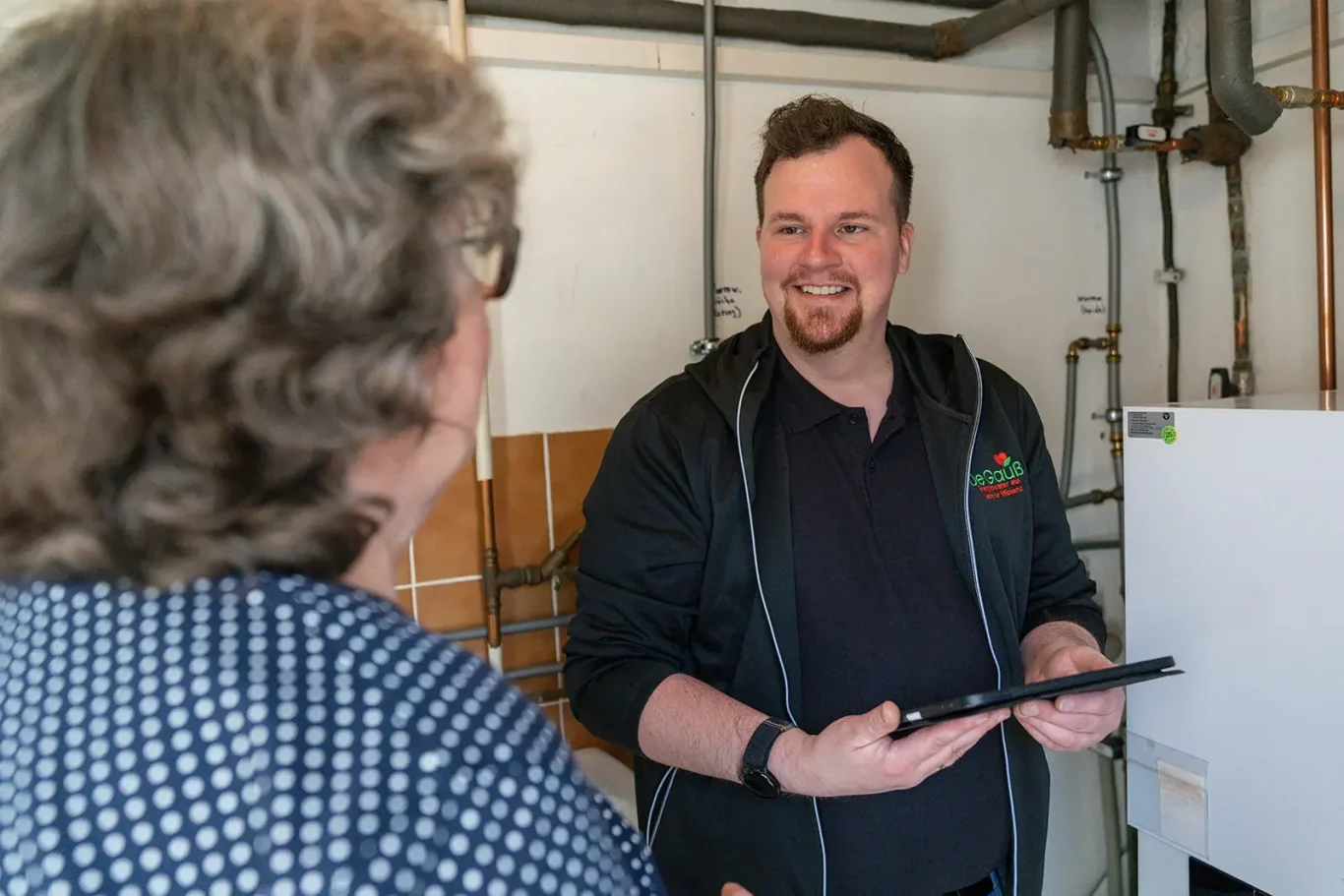 Energieberater Dennis Gauß berät eine Kundin im Heizungskeller zum Thema Wärmepumpen-Eignung und nutzt ein Tablet zur Datenerfassung.